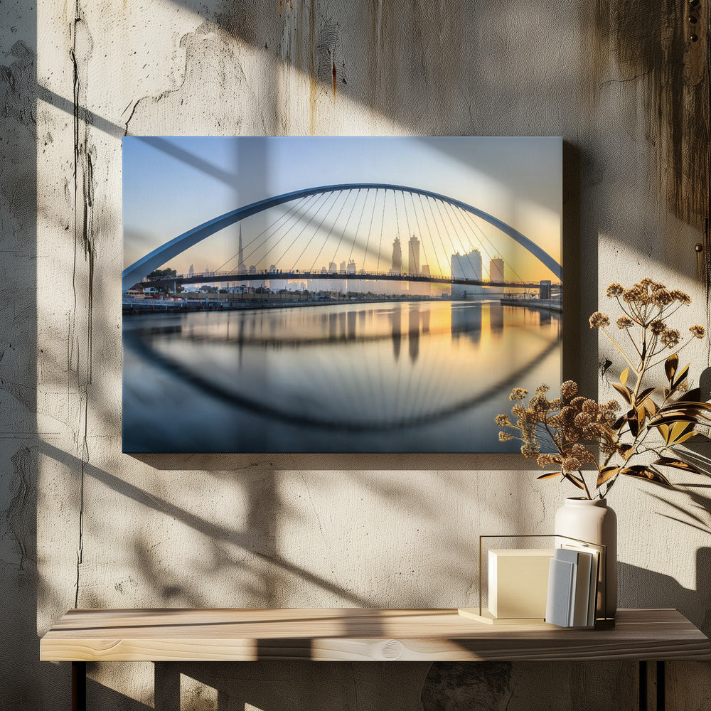 A framed photograph of the Dubai Water Canal Bridge at sunrise, with its large arch spanning across the water. The Dubai skyline, including the Burj Khalifa, is visible in the background, silhouetted against a golden and blue sky. The bridge and buildings create a perfect reflection on the calm surface of the water. Wall Art