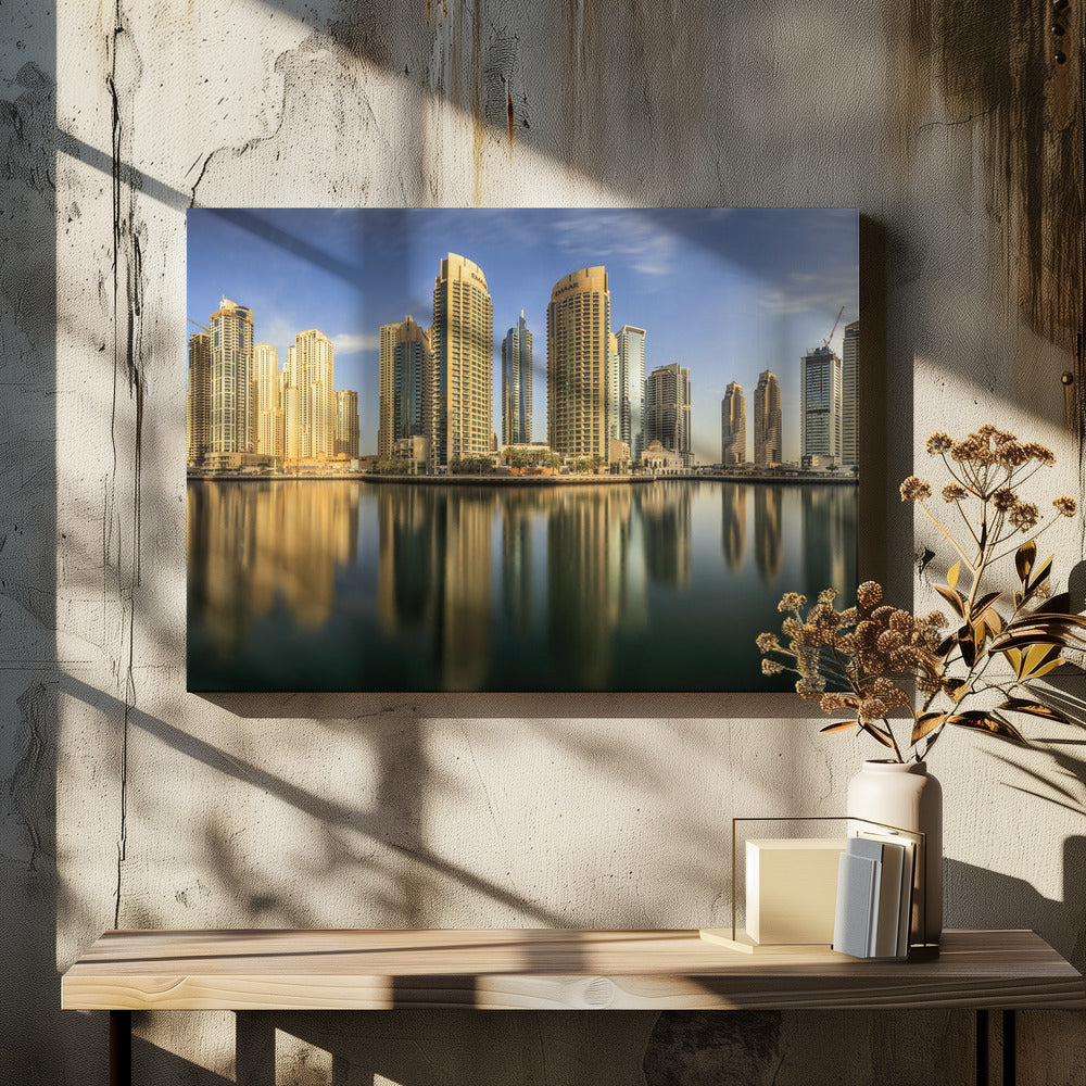 A panoramic photograph of the Dubai Marina skyline with its golden skyscrapers reflecting in the calm, dark water below, all under a clear blue sky and framed in silver. Wall Art