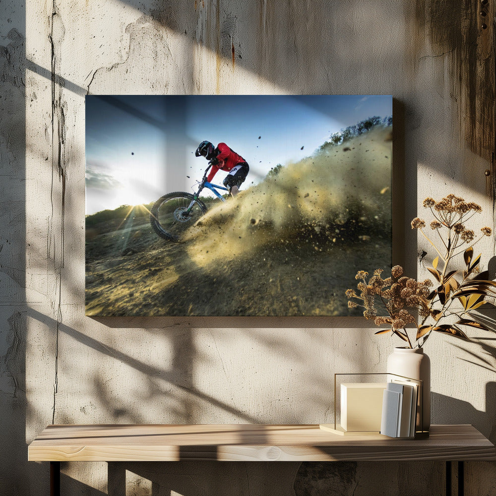 A mountain biker wearing a red jersey and helmet speeds down a steep, dusty trail, kicking up a large cloud of dirt. The sun is setting in the background, creating a lens flare against a clear blue sky. The image is captured from a low angle, emphasizing the action and speed, and is presented within a silver frame. Artwork