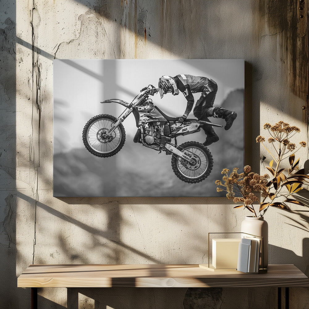 A dynamic black and white photograph of a motocross rider in mid-air, performing a jump. The rider, wearing full protective gear and a helmet, is leaning forward over the handlebars of the dirt bike. The bike is suspended against a blurry, light gray background, emphasizing the action and height of the jump. The image is enclosed in a silver frame. Wall Art