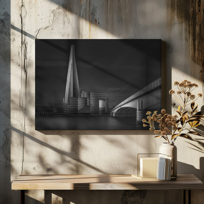 A dramatic black and white photograph of the London skyline featuring The Shard skyscraper and London Bridge. The image is shot at dusk or night, with The Shard brightly lit against a dark, moody sky. The River Thames in the foreground is calm and reflective, captured with a long exposure. Artwork