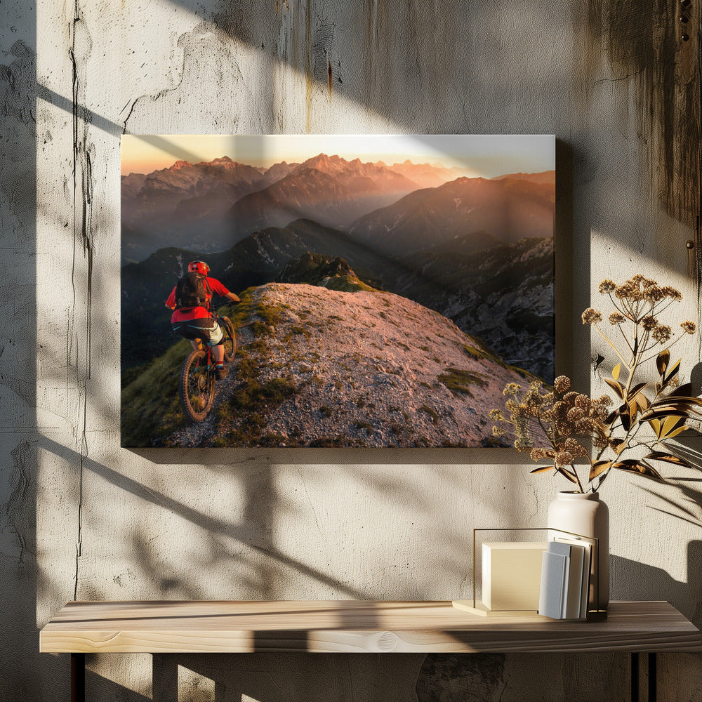 A mountain biker wearing a red shirt and helmet rides along a narrow, rocky mountain ridge at sunset. The background features a vast landscape of layered mountain ranges glowing in the warm, golden light of the setting sun. Poster