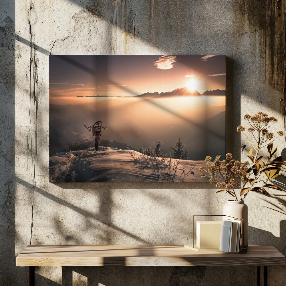 A lone person stands on a snowy mountain peak at sunrise, carrying a bicycle over their shoulder. They look out over a valley filled with a sea of clouds, with distant mountain ranges silhouetted against the bright, golden sun. Artwork