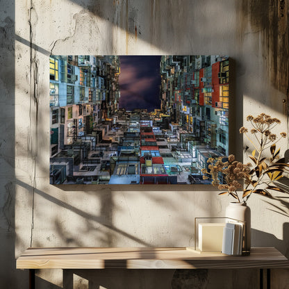 A low-angle nighttime view looking up from a courtyard surrounded by extremely dense, stacked apartment buildings with many windows lit in yellow, blue, and white, revealing a small patch of dark, cloudy sky. The image is presented within a brushed silver frame. Artwork