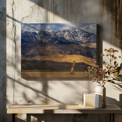 A framed photograph of a land sailor with a bright orange and blue sail on a vast, flat desert floor. In the background, majestic snow-dusted mountains rise against a cloudy blue sky. Artwork
