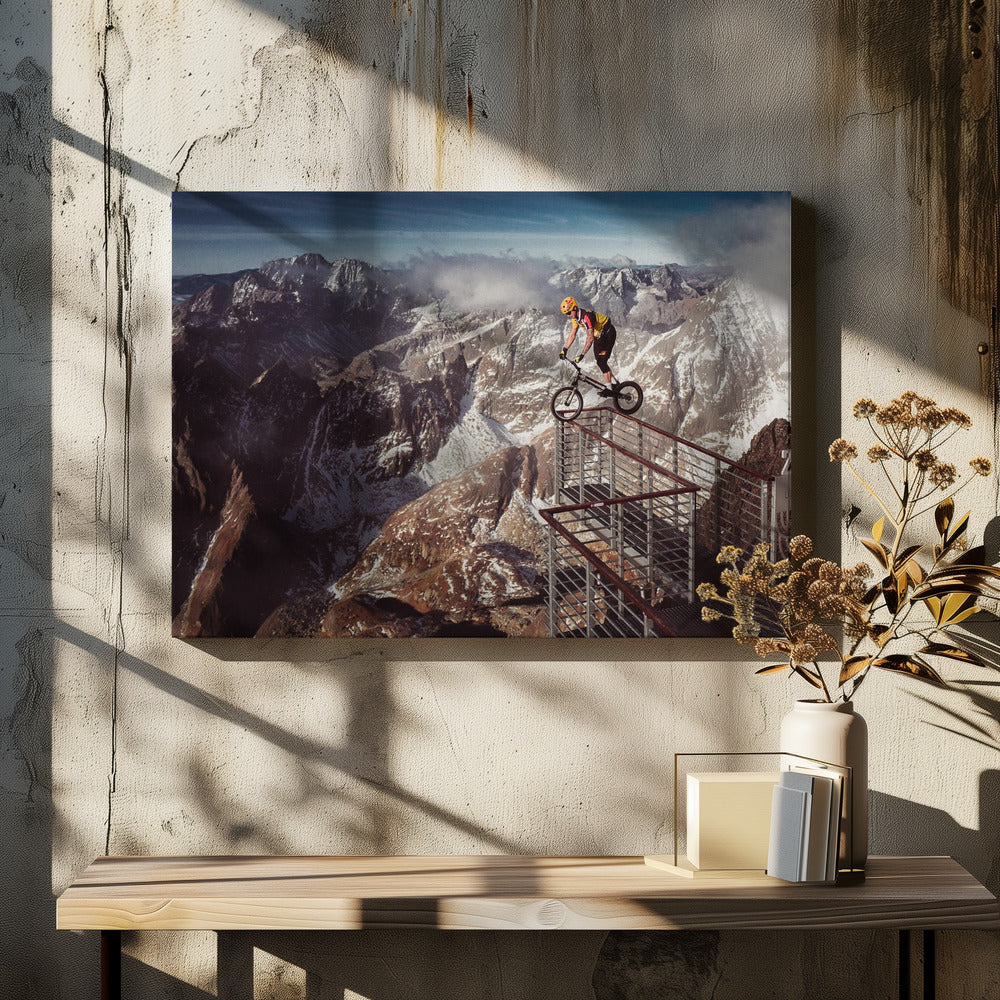 A trials cyclist wearing a yellow helmet balances precariously on the railing of a metal walkway high in the mountains, with a dramatic background of snow-dusted peaks and a blue sky. Poster