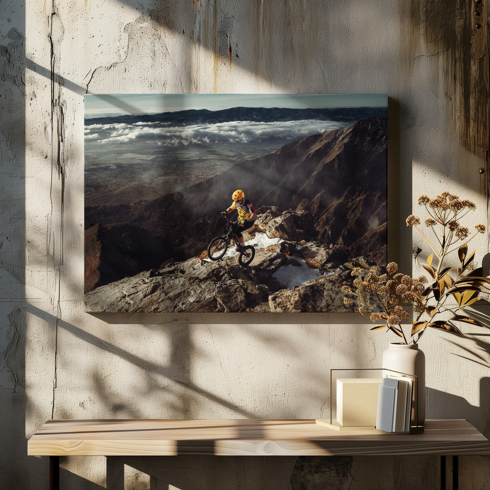 A mountain biker in a yellow helmet and jersey navigates a treacherous, rocky mountain ridge. Below, a vast valley is filled with a sea of clouds, with distant mountain ranges visible on the horizon. Print