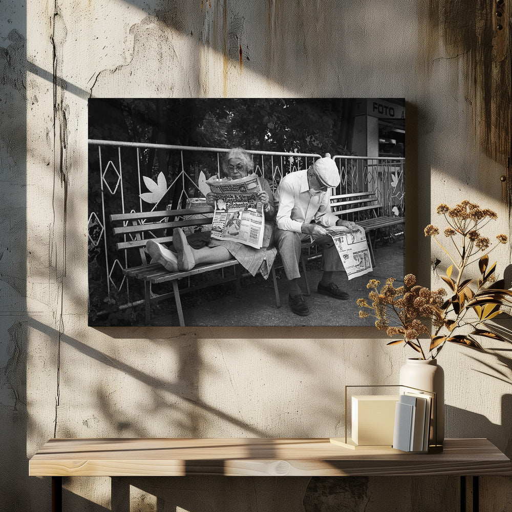 A framed black and white photograph of an elderly couple sitting on a park bench, both deeply absorbed in reading their newspapers. The woman on the left has her feet propped up on the bench, while the man on the right sits next to her, looking down at his paper. Poster