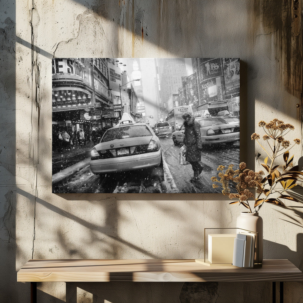 A black and white photograph of a bustling New York City street during a snowstorm. A person in a heavy winter coat crosses the street in front of a line of yellow cabs, with the bright lights of Times Square billboards glowing softly in the background through the falling snow. Print