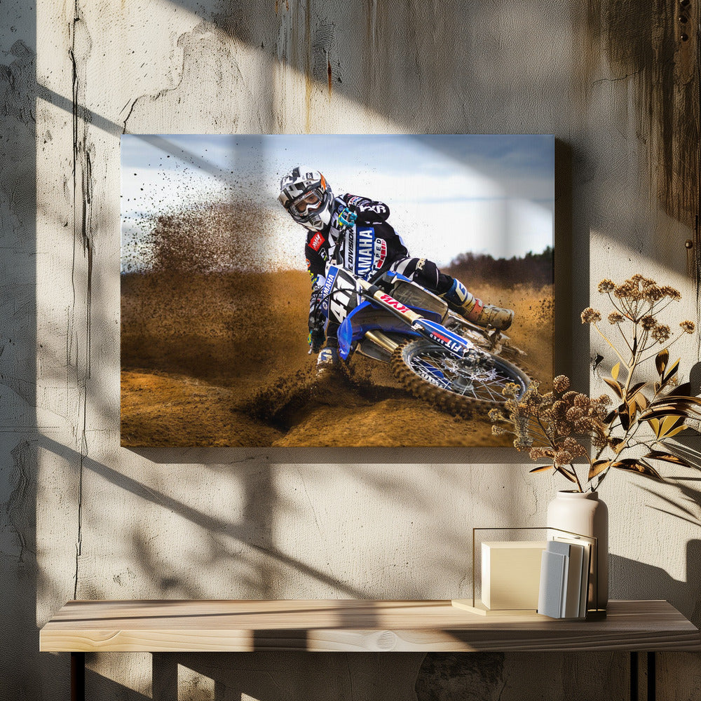A motocross rider in full gear leans sharply into a turn on a dirt track, kicking up a large spray of mud and dirt from their blue and white Yamaha motorcycle. Artwork