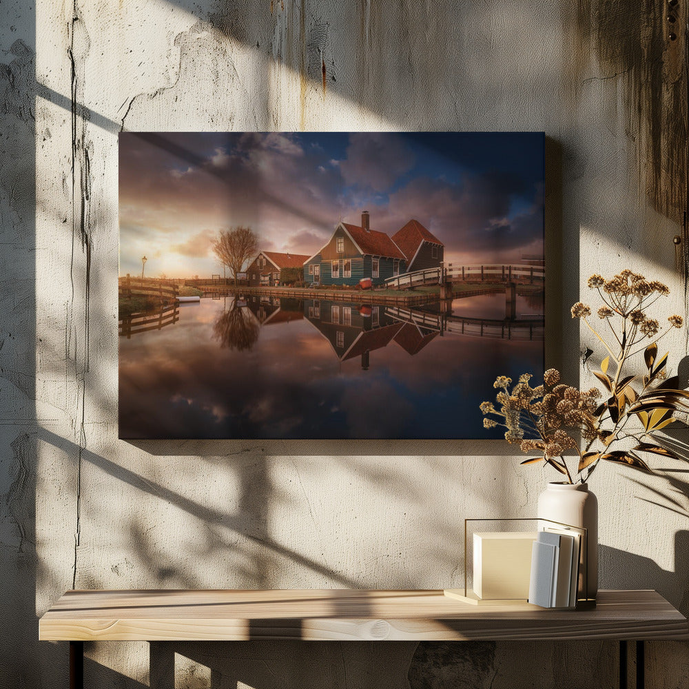 A picturesque landscape of traditional Dutch houses with teal walls and red roofs reflected perfectly in the calm water of a canal at sunset, under a dramatic, cloudy sky. A small wooden bridge crosses the canal, and the scene is enclosed in a silver frame. Artwork
