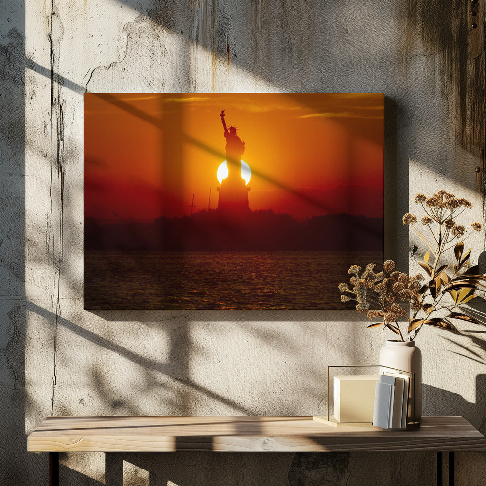 A powerful silhouette of the Statue of Liberty against a fiery orange and red sunset. The sun is perfectly positioned behind the statue's base, creating a brilliant, glowing orb of light. The dark waters in the foreground reflect the vibrant colors of the sky. The photograph is presented within a silver-colored frame. Artwork
