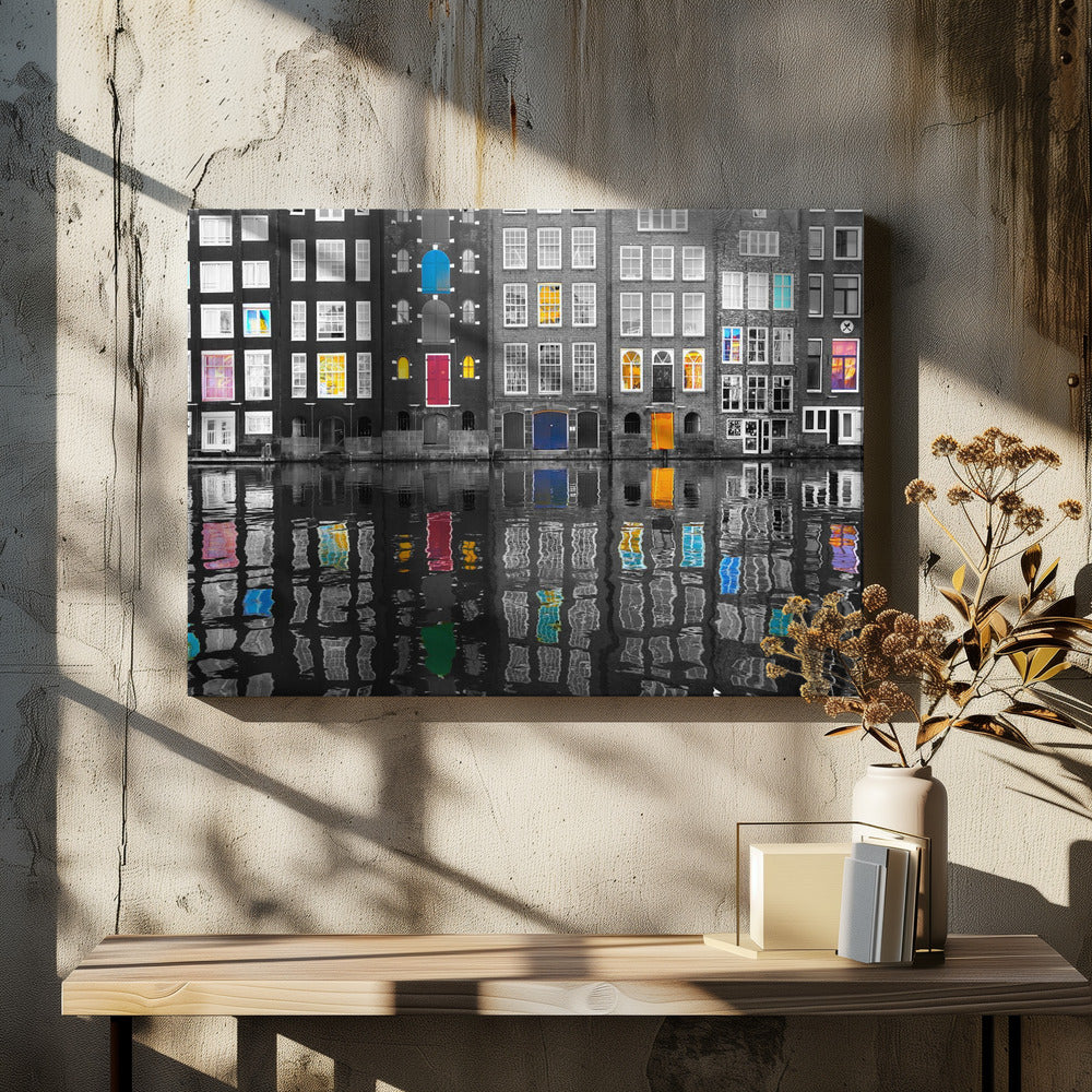 A framed black and white photograph of canal houses in Amsterdam, with selective coloring highlighting windows and doors in vibrant shades of red, blue, yellow, and pink. The colorful architectural details are mirrored in the rippling water of the canal below. Artwork