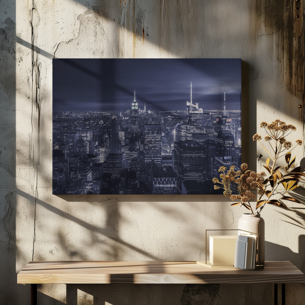 A dramatic nighttime photograph of the New York City skyline, viewed from a high vantage point. The cityscape is a sea of illuminated skyscrapers under a dark, moody sky, with the entire image cast in a cool blue and white monochrome tone and presented in a silver frame. Artwork