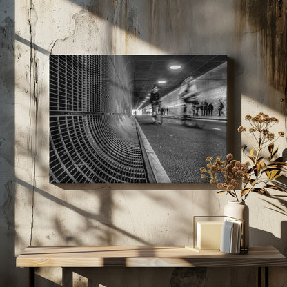 A dynamic black and white photograph from inside a modern tunnel. On the left, a curved metal grate wall creates a strong pattern, while on the right, cyclists are captured in motion blur on a paved path. Pedestrians are visible in the background, walking through the illuminated passage. Poster