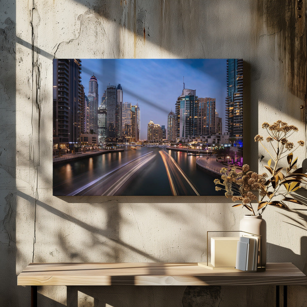 A long-exposure photograph of the Dubai Marina at dusk, framed in silver. Illuminated skyscrapers line a busy waterway, with light trails from moving boats creating long, bright streaks on the water. The sky is a deep twilight blue, and the city's lights cast a warm glow. Decor