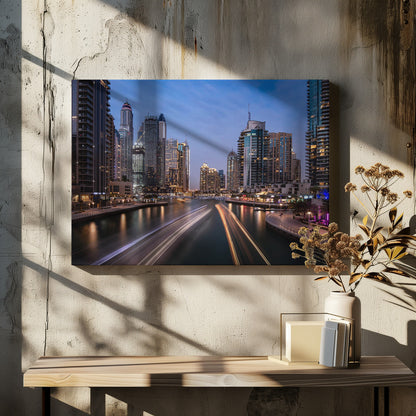 A long-exposure photograph of the Dubai Marina at dusk, framed in silver. Illuminated skyscrapers line a busy waterway, with light trails from moving boats creating long, bright streaks on the water. The sky is a deep twilight blue, and the city's lights cast a warm glow. Decor