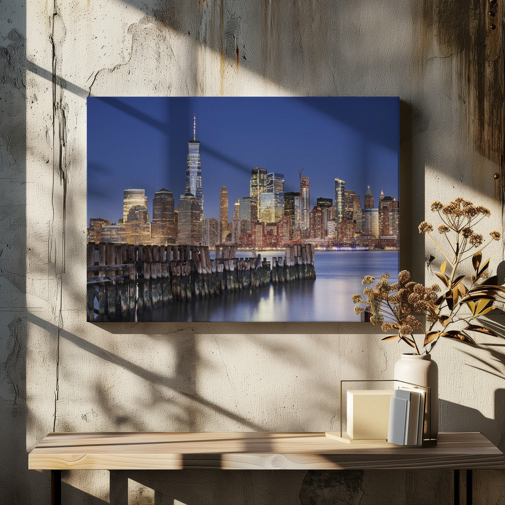 A framed photograph of the Lower Manhattan skyline at dusk. The One World Trade Center and other skyscrapers are brightly lit against a deep blue sky. In the foreground, an old wooden pier extends into the water, which reflects the city lights. Wall Art