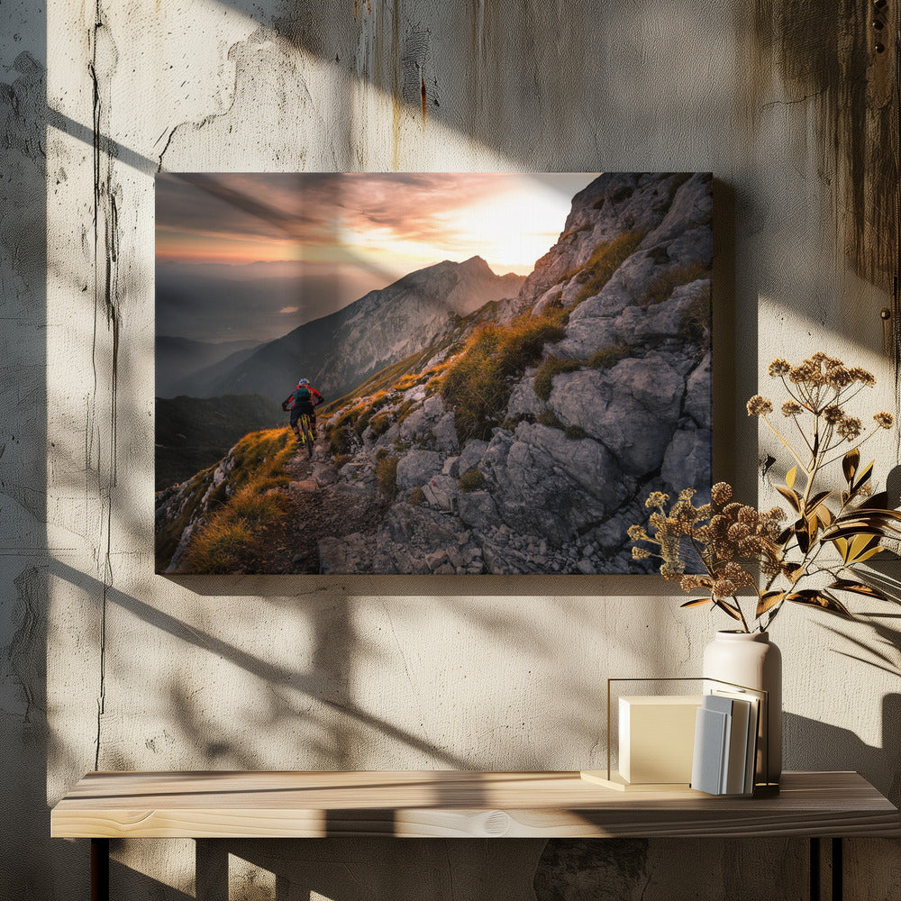 A mountain biker from behind rides along a narrow, rocky trail on a steep mountain ridge during a dramatic golden sunset. In the background, layers of mountains fade into the distance under a warm, glowing sky. Artwork