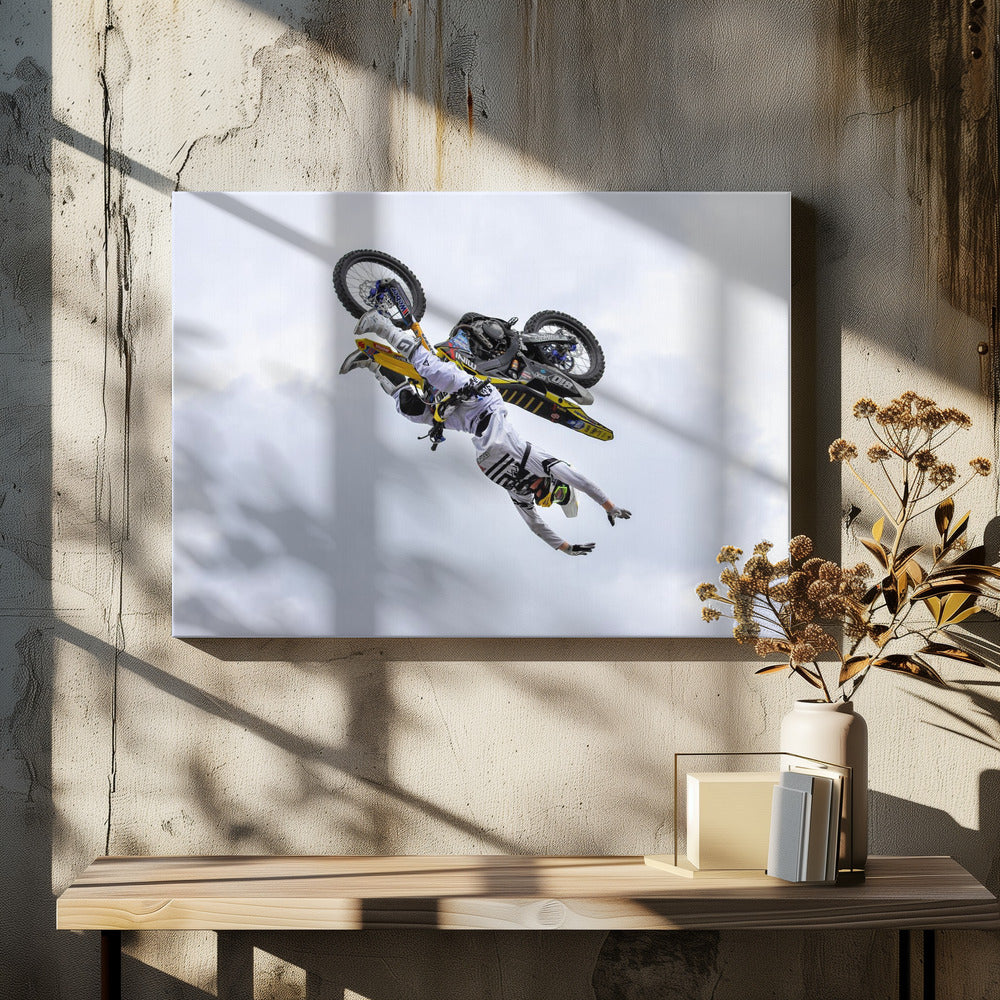 A framed action shot of a freestyle motocross rider in mid-air, performing an upside-down trick against a cloudy sky, with their body hanging below the yellow and black dirt bike. Artwork