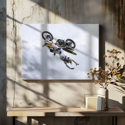 A framed action shot of a freestyle motocross rider in mid-air, performing an upside-down trick against a cloudy sky, with their body hanging below the yellow and black dirt bike. Artwork
