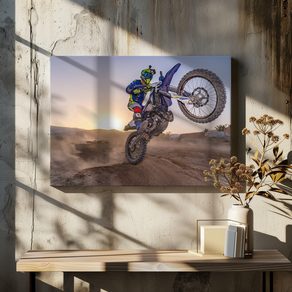 A motocross rider in blue and yellow gear is captured mid-air on a blue dirt bike, performing a jump against a dusty landscape with a setting sun in the background. The image is enclosed in a silver frame. Wall Art