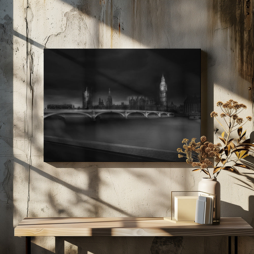 A moody, atmospheric black and white photograph of the Houses of Parliament and Big Ben in London, viewed from across the River Thames. The iconic buildings are softly illuminated against a dark, dramatic sky, with Westminster Bridge arching over the dark, smooth water in the foreground. The image is presented within a silver-colored frame. Artwork
