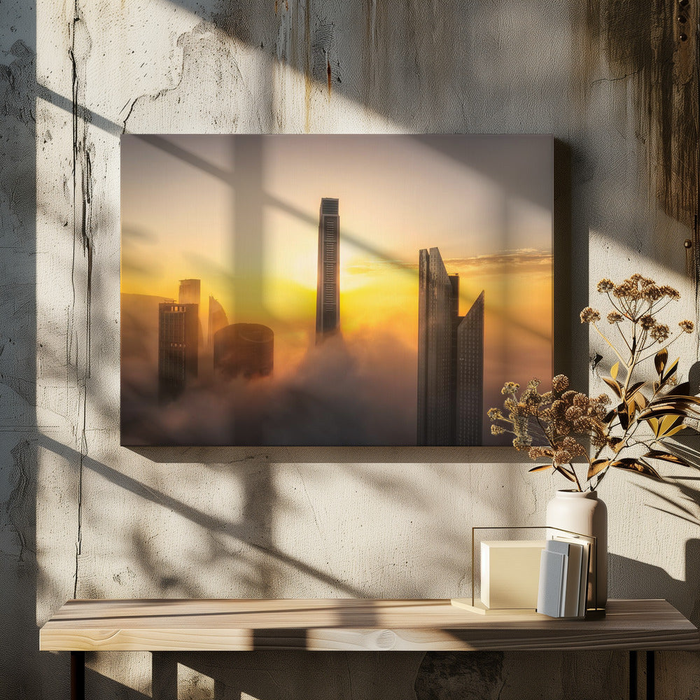 A framed photograph of a modern city skyline with skyscrapers emerging from a thick layer of clouds at sunrise, creating a dramatic and ethereal scene against a golden sky. Wall Art