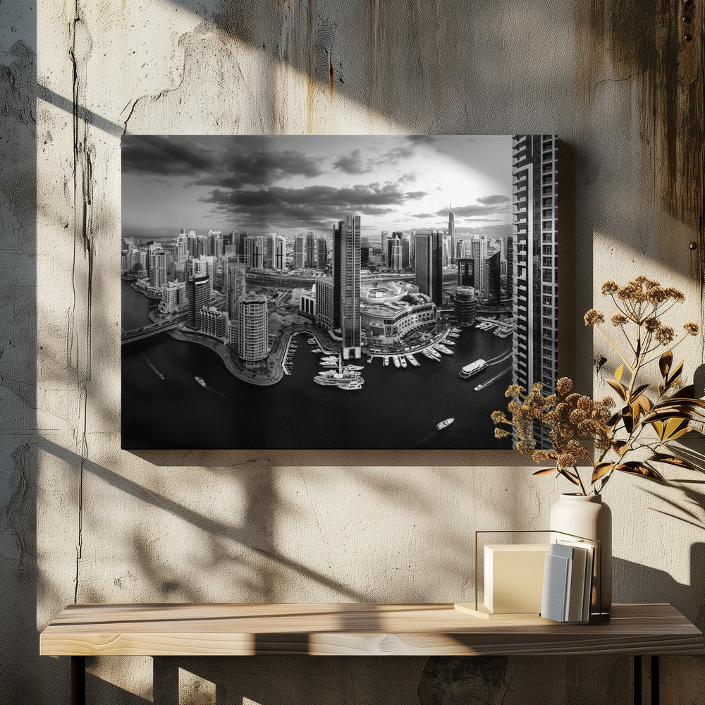 A framed, black and white panoramic photograph of the Dubai Marina taken from a high vantage point. The image captures the dense cityscape with numerous modern skyscrapers surrounding a winding waterway filled with docked yachts and moving boats. The sky is filled with dramatic, dark clouds, adding to the moody atmosphere of the urban landscape. Decor