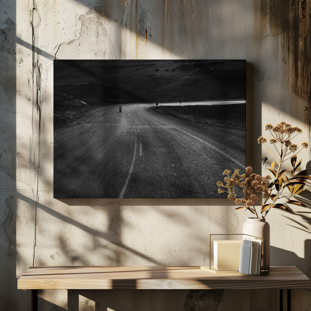 A moody, black and white photograph capturing a lone motorcyclist from behind as they navigate a curving road through a dark, hilly terrain, framed in silver. Decor
