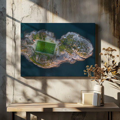 An aerial view of a vibrant green soccer field situated on a small, rocky island surrounded by dark blue water. The photograph, taken from directly above, captures the unique and remote location of the sports facility, with small buildings and wooden structures dotting the island's terrain. The entire scene is enclosed within a silver frame. Print