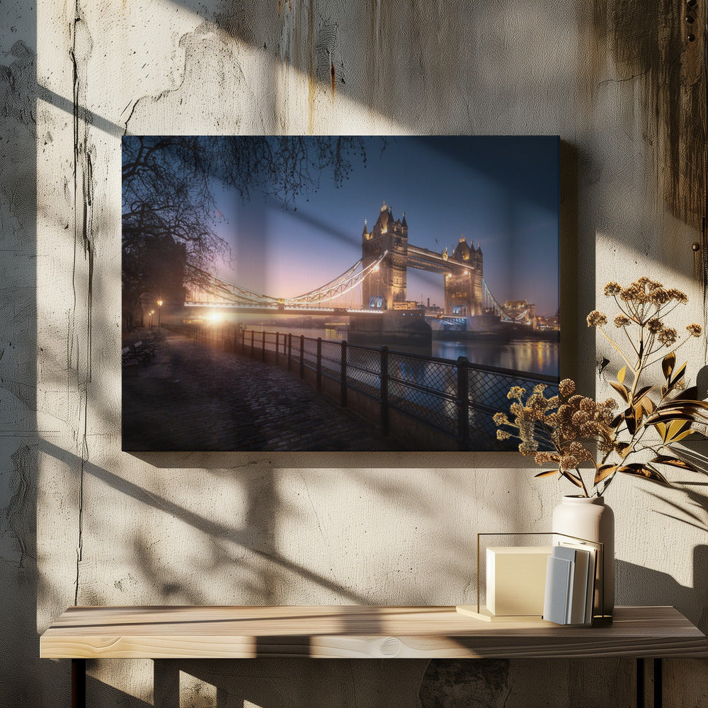A scenic photograph of London's Tower Bridge at sunrise, its lights glowing warmly against a deep blue and orange sky. In the foreground, a cobblestone path follows the River Thames, leading the eye towards the iconic bridge and the rising sun. Poster