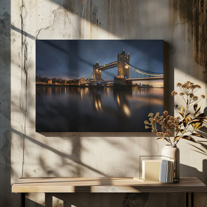 A long-exposure photograph of London's Tower Bridge at dusk, brilliantly illuminated with golden lights that cast shimmering reflections on the calm River Thames. The sky is a deep, dark blue, and the city skyline glows in the background, all enclosed within a silver frame. Print