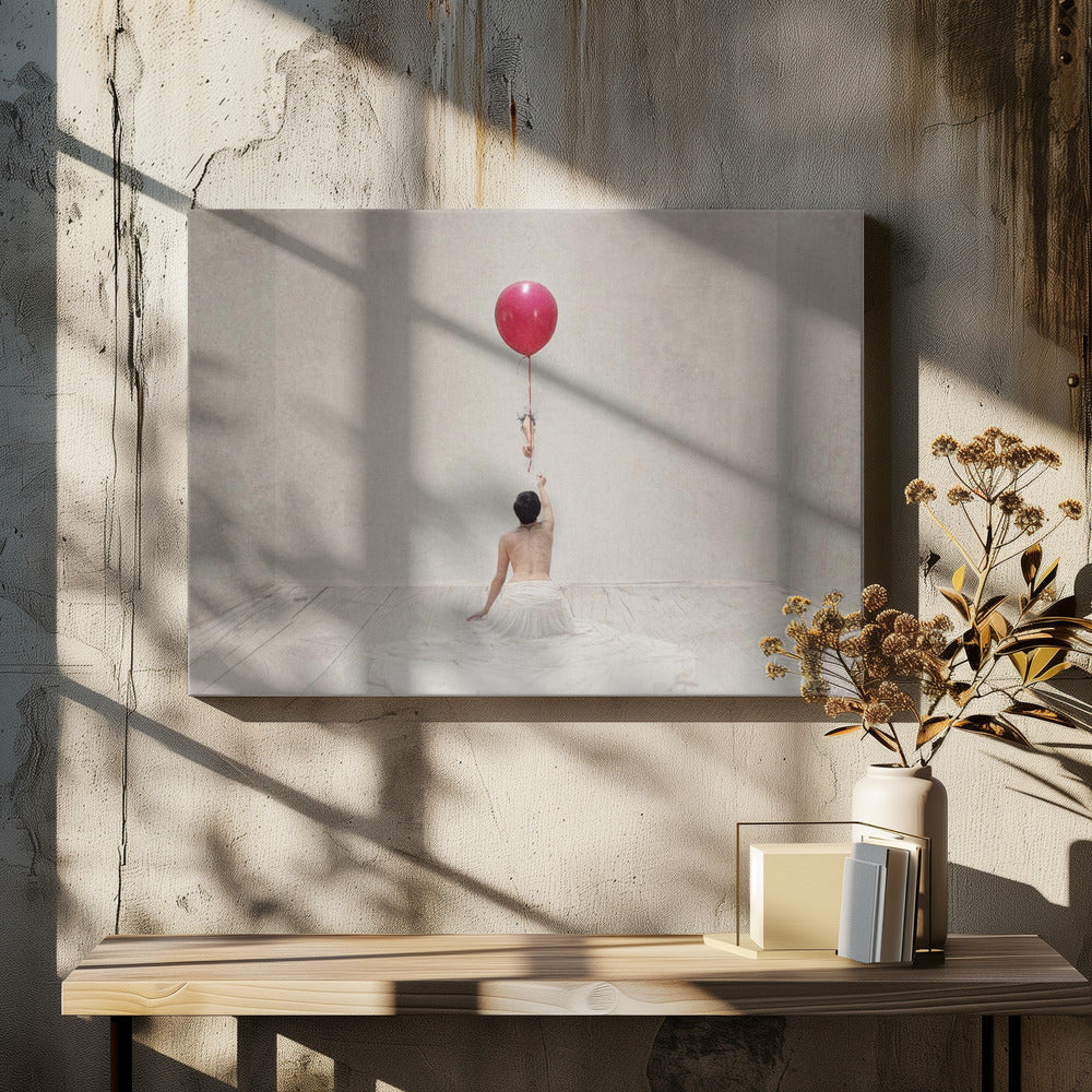 A surreal fine art photograph of a person with a bare back and a long white skirt, sitting on the floor of a minimalist grey room. They are reaching up towards a small figure hanging from the string of a large, bright red balloon that floats towards the ceiling. The image is enclosed in a silver frame. Artwork