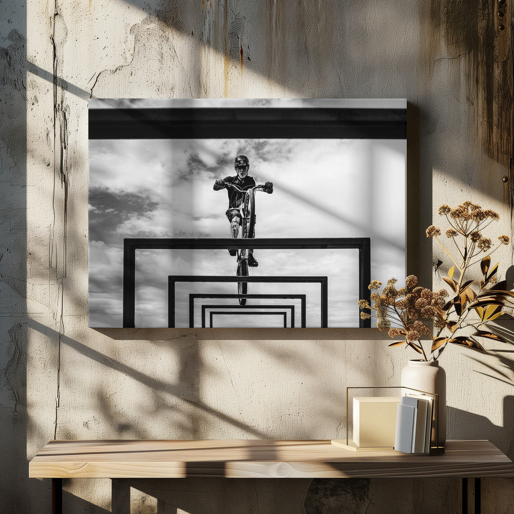 A striking black and white photograph taken from a low angle, showing a cyclist in full gear balancing on a mountain bike. The cyclist is positioned centrally above a series of black rectangular frames that create a receding, tunnel-like visual effect against a cloudy sky. The entire image is presented within a silver picture frame. Artwork
