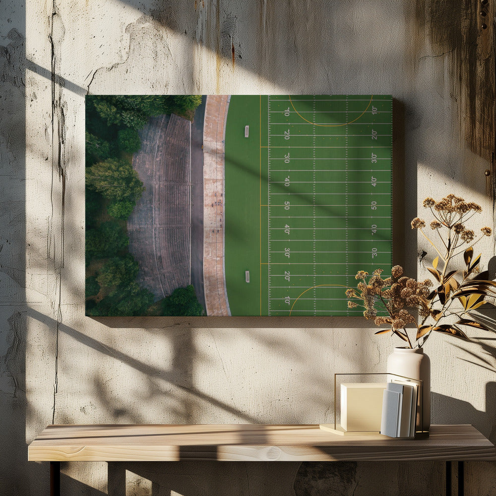 An aerial top-down photograph of an American football field beside empty, weathered concrete stadium bleachers. The vibrant green field is marked with white yard lines, and the gray stands are nestled amongst lush green trees. The entire scene is enclosed in a silver frame. Artwork