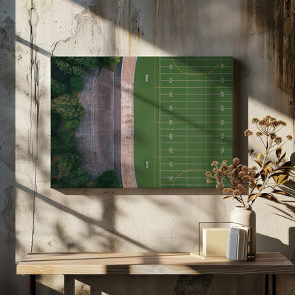 An aerial top-down photograph of an American football field beside empty, weathered concrete stadium bleachers. The vibrant green field is marked with white yard lines, and the gray stands are nestled amongst lush green trees. The entire scene is enclosed in a silver frame. Artwork