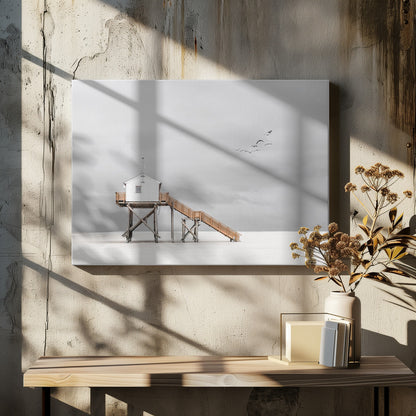 A minimalist, desaturated photograph of a small white cabin on tall wooden stilts on a vast, empty beach. A long wooden staircase leads up to the cabin. The sky is a flat, overcast grey, and a flock of dark birds is visible flying in the distance. Print