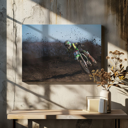 A motocross rider in green and black gear leans hard into a turn on a dirt track, kicking up a large spray of mud against a clear blue sky. Decor