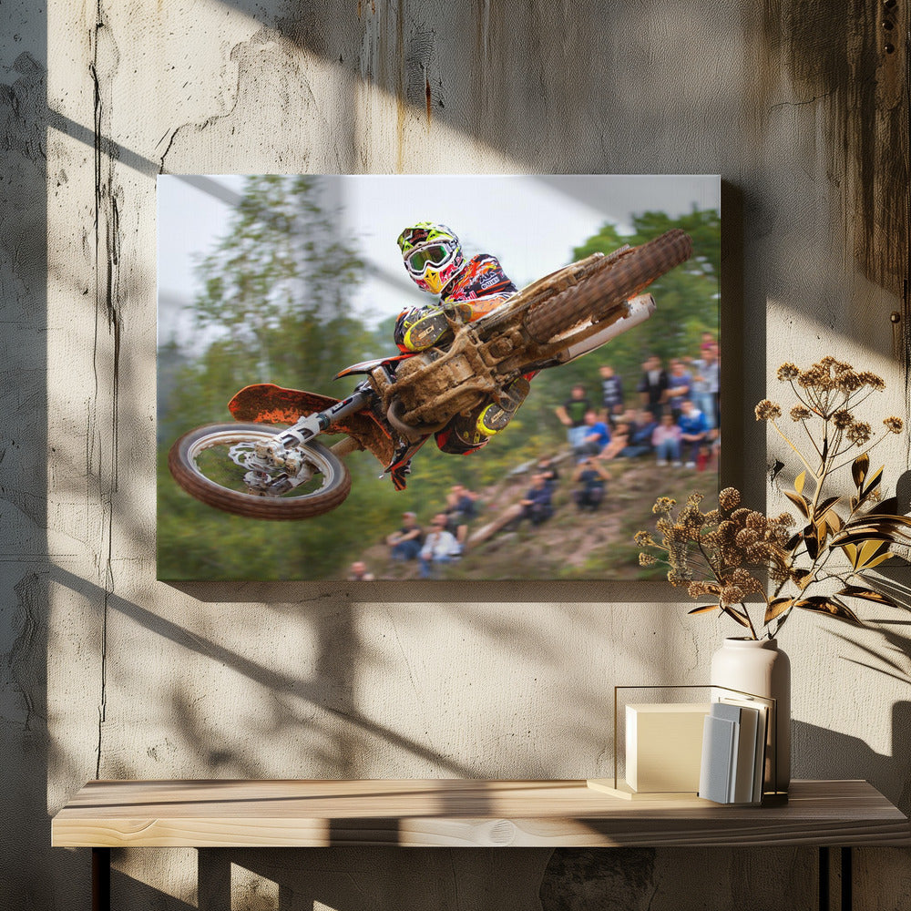 A dynamic action shot of a motocross racer in mid-air, leaning into a turn with their muddy orange dirt bike. The rider wears a colorful helmet and racing gear, with a blurred background of green trees and spectators watching from a dirt hill. The photo is enclosed in a silver frame. Decor
