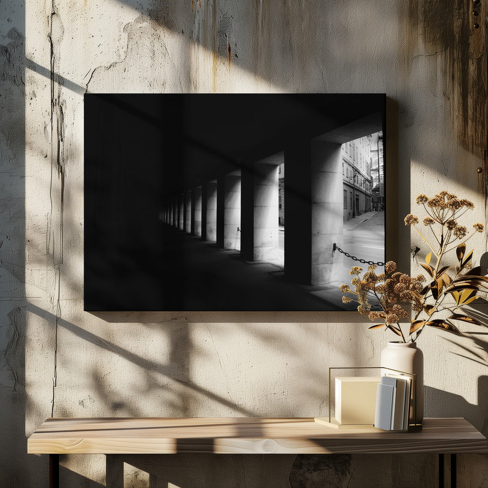 A high-contrast, black and white photograph from the perspective of a dark, covered walkway lined with a row of thick pillars. Light from an adjacent street highlights the pillars, creating a dramatic play of light and shadow and a strong sense of depth. Print