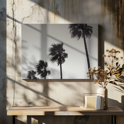 A framed black and white photograph of four silhouetted palm trees of increasing height, arranged diagonally against a plain, light grey sky. The palm fronds are blurred, suggesting they are blowing in the wind. Decor