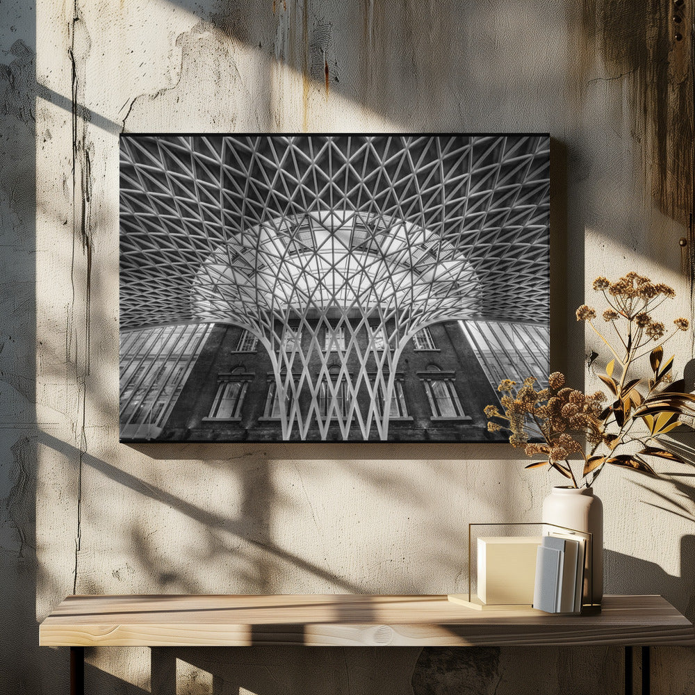 A framed, black and white photograph of the Western Concourse at King's Cross Station, London. The view is from a low angle, looking up at the intricate, white, funnel-like lattice roof structure as it spreads out across the ceiling, contrasting with the classic brick facade of the building behind it. Decor