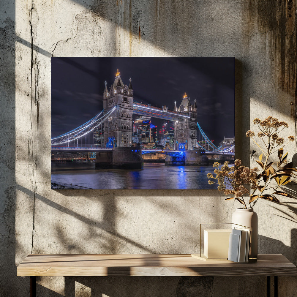 A vibrant night photograph of the iconic Tower Bridge in London, illuminated with bright white and electric blue lights. The dark River Thames in the foreground reflects the bridge and the lit-up modern city skyline in the background, all under a dark, cloudy sky. The image is enclosed in a silver frame. Decor