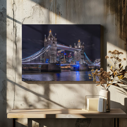 A vibrant night photograph of the iconic Tower Bridge in London, illuminated with bright white and electric blue lights. The dark River Thames in the foreground reflects the bridge and the lit-up modern city skyline in the background, all under a dark, cloudy sky. The image is enclosed in a silver frame. Decor