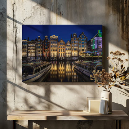 A vibrant night-time photograph of a canal in Amsterdam, flanked by two tour boats in the foreground. Traditional Dutch canal houses are brightly lit, casting warm yellow reflections on the dark water under a deep blue sky. Print
