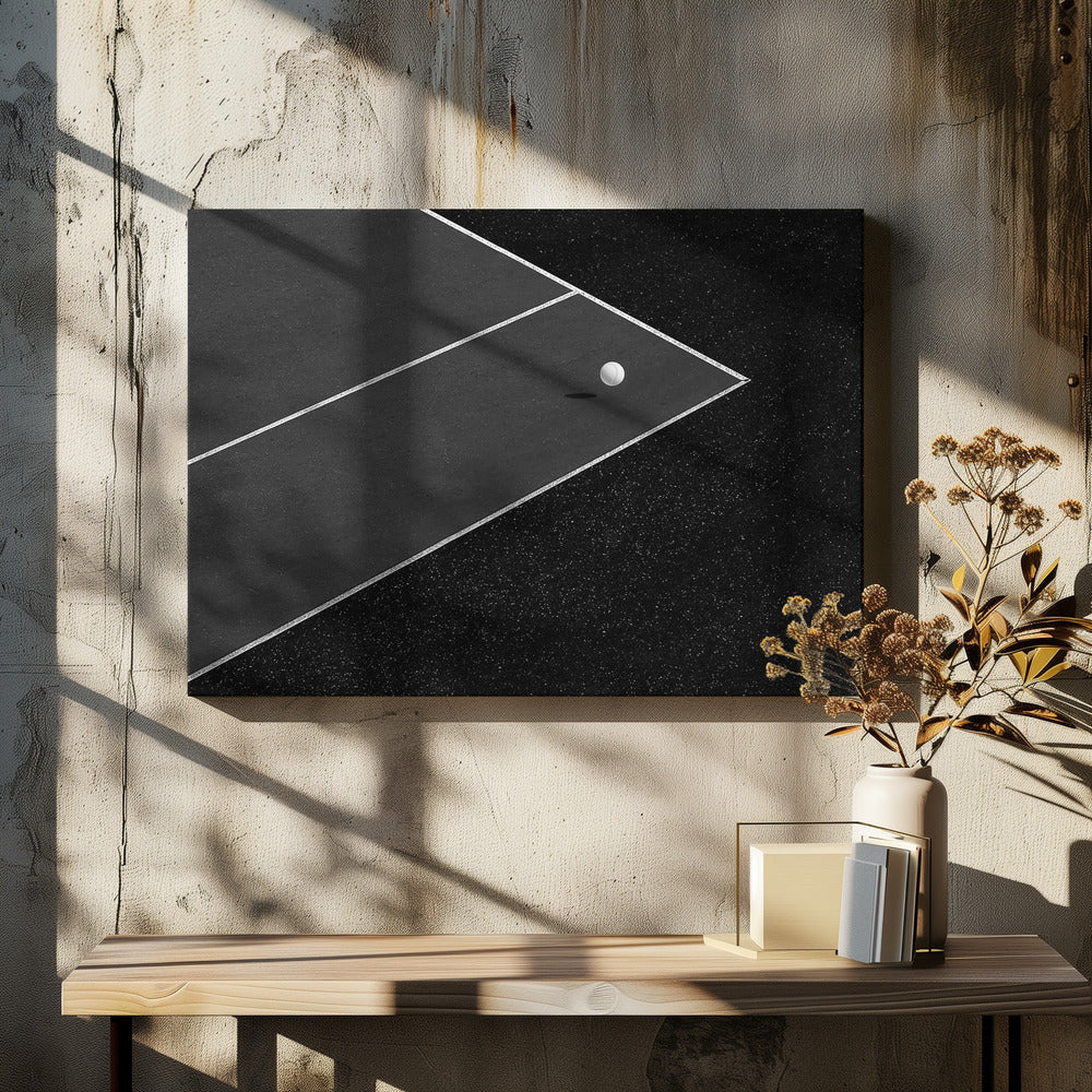 A minimalist black and white overhead photograph of a white ball resting in the corner of a dark tennis or paddle court. The composition focuses on the strong geometric angles created by the white boundary lines against the dark, textured asphalt, all within a silver frame. Artwork