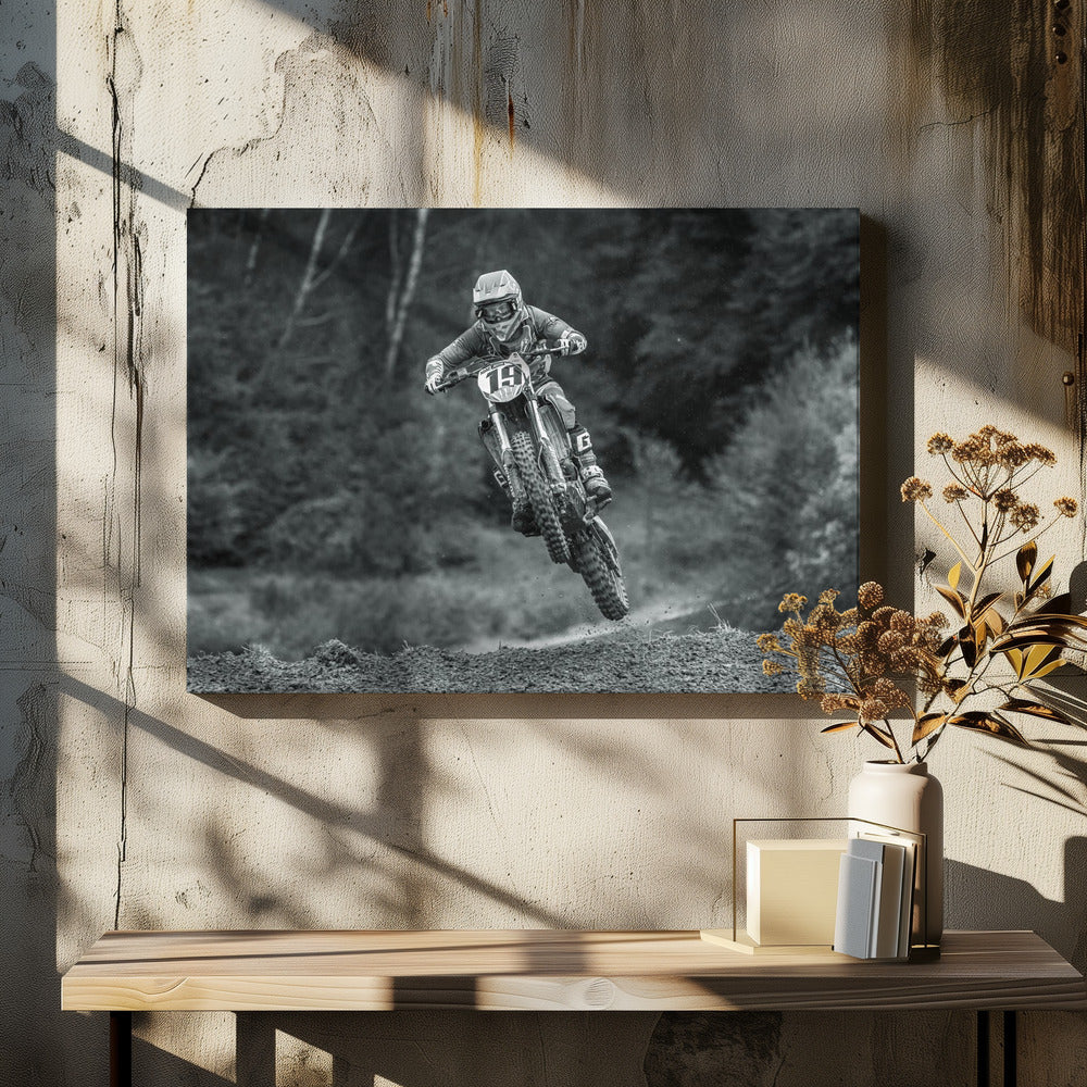 A dynamic black and white photograph of a motocross racer in mid-air, jumping over a dirt mound. The rider, wearing full protective gear and a helmet, is on a dirt bike with the number 19 plate. The background is a soft-focus forest, and the entire image is enclosed in a silver frame. Poster