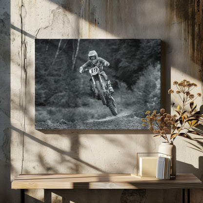 A dynamic black and white photograph of a motocross racer in mid-air, jumping over a dirt mound. The rider, wearing full protective gear and a helmet, is on a dirt bike with the number 19 plate. The background is a soft-focus forest, and the entire image is enclosed in a silver frame. Poster