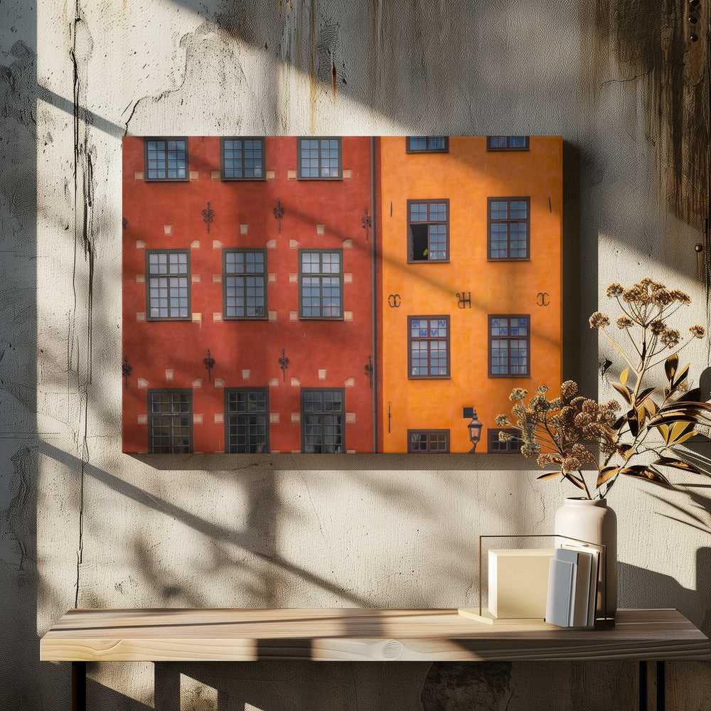 A framed close-up photograph of two adjacent, colorful building facades. The building on the left is a deep, rustic red, while the one on the right is a vibrant orange. Both feature numerous multi-paned windows with dark frames. Artwork